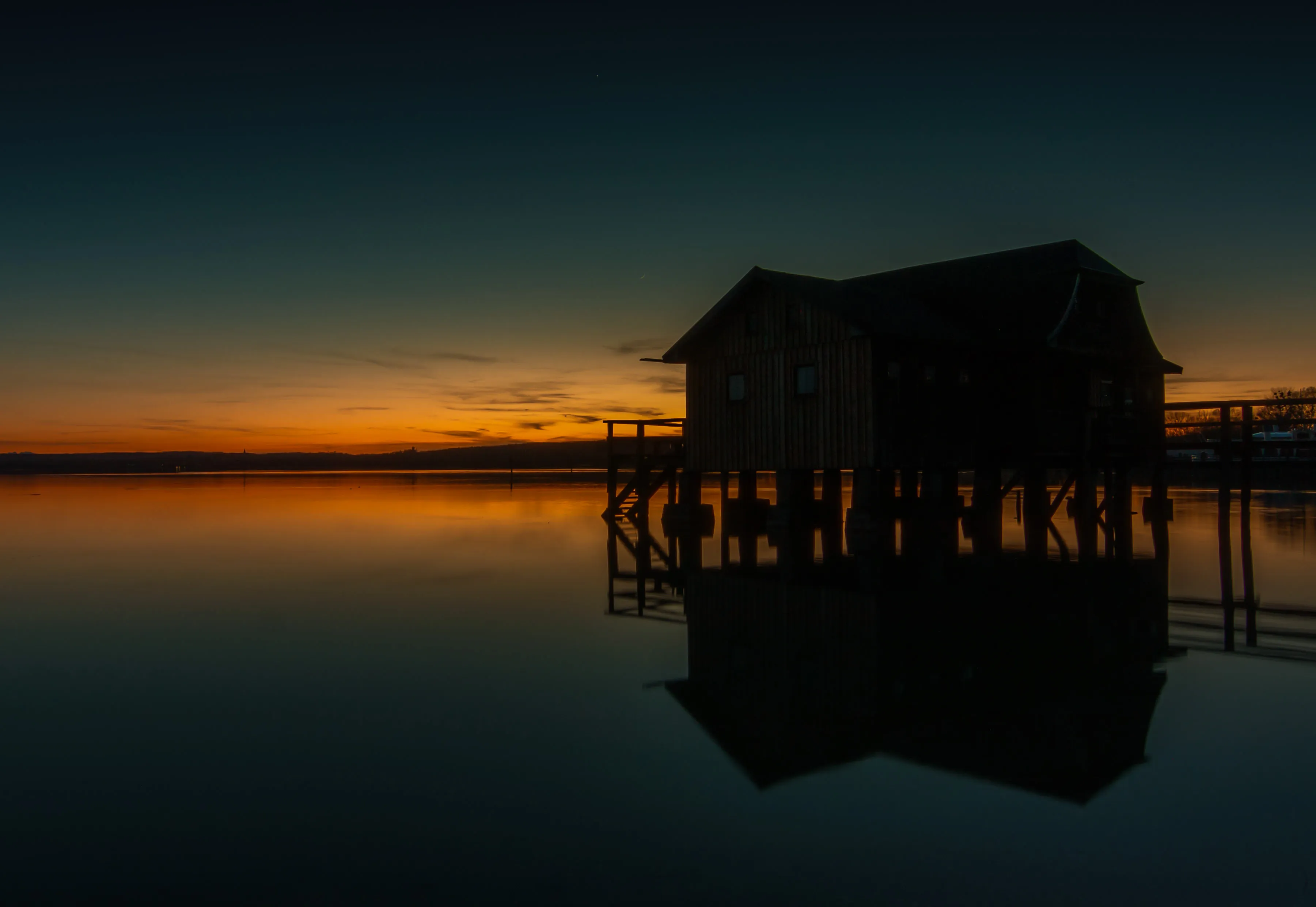 Ein Bootshaus im Ammersee zur Dämmerung, der Himmel spiegelt sich im Wasser