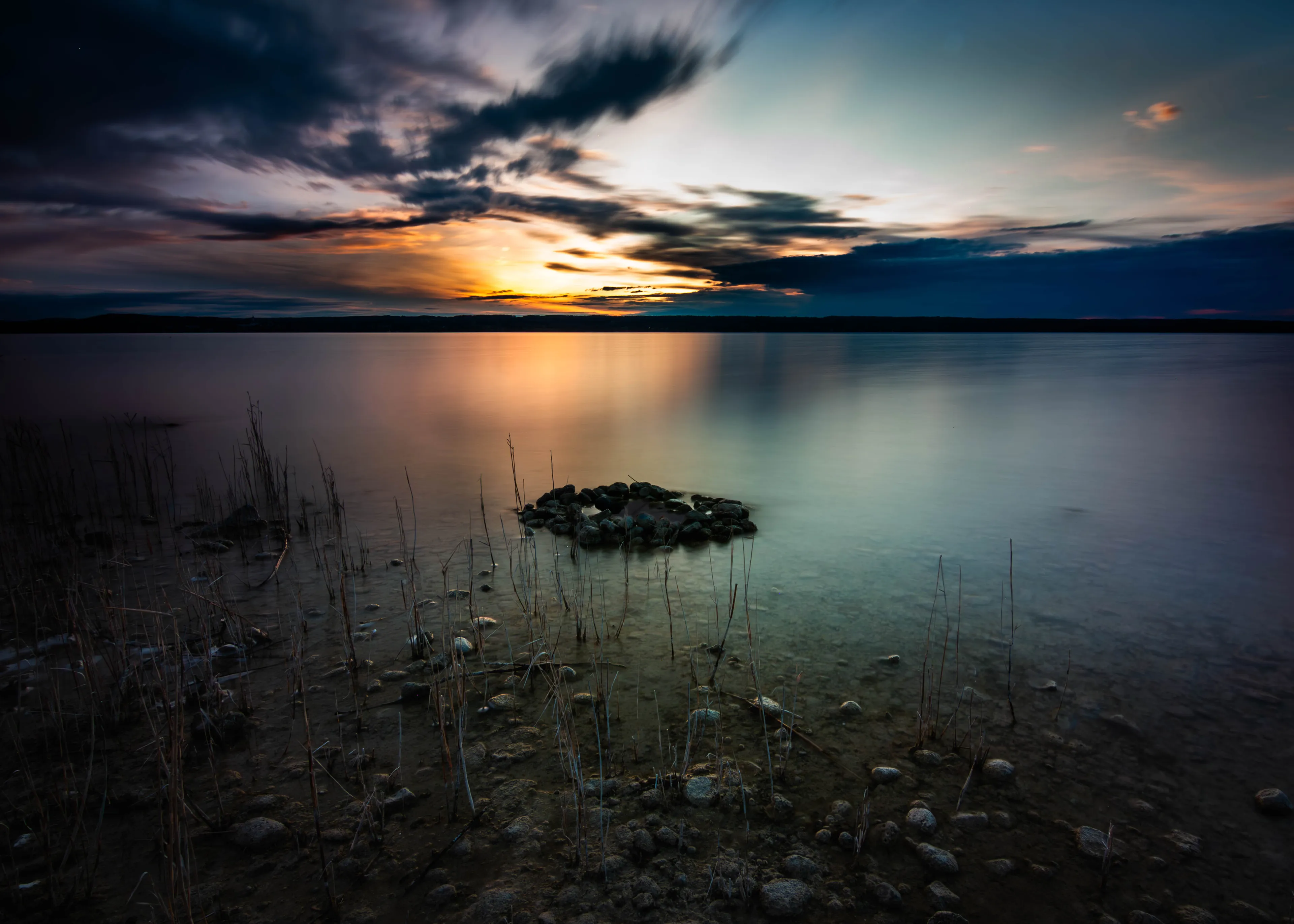 Sonnenuntergang über dem Ammersee, im Vordergrund Schilf und Steine im flachen Wasser