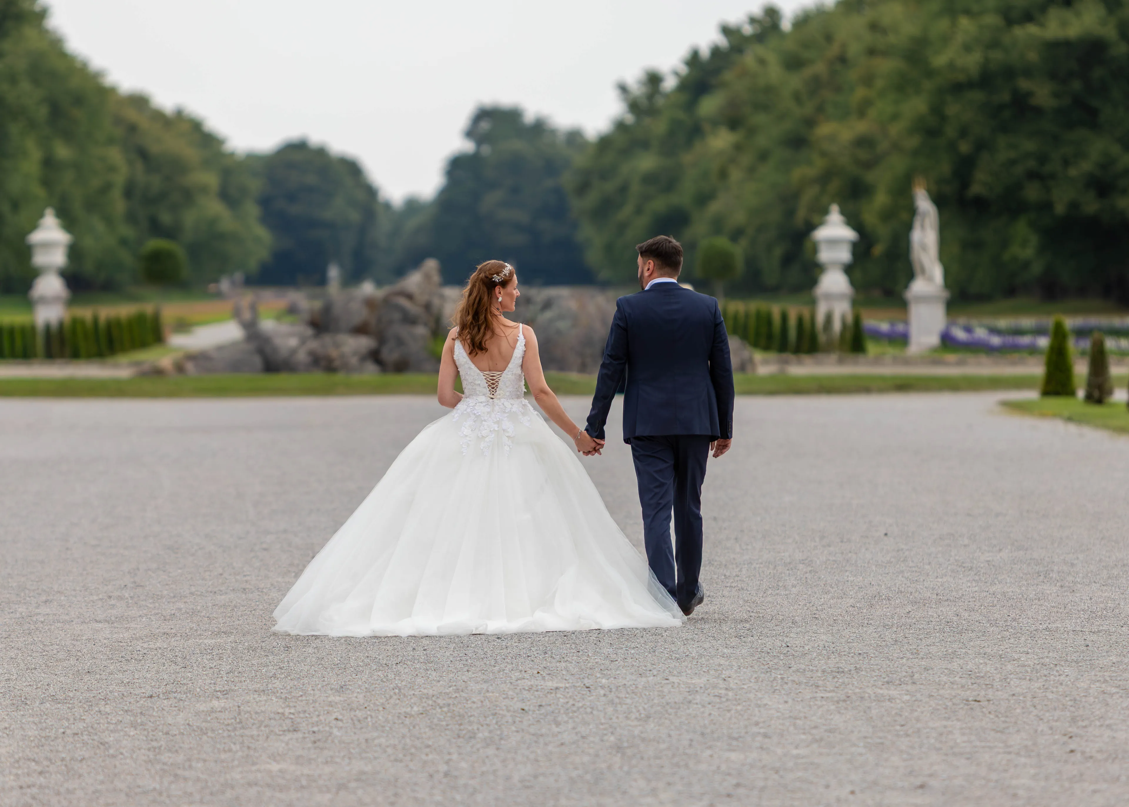 Brautpaar geht Hand in Hand über den Kiesweg im Schlosspark Nymphenburg, links und rechts Statuen und geschnittene Hecken