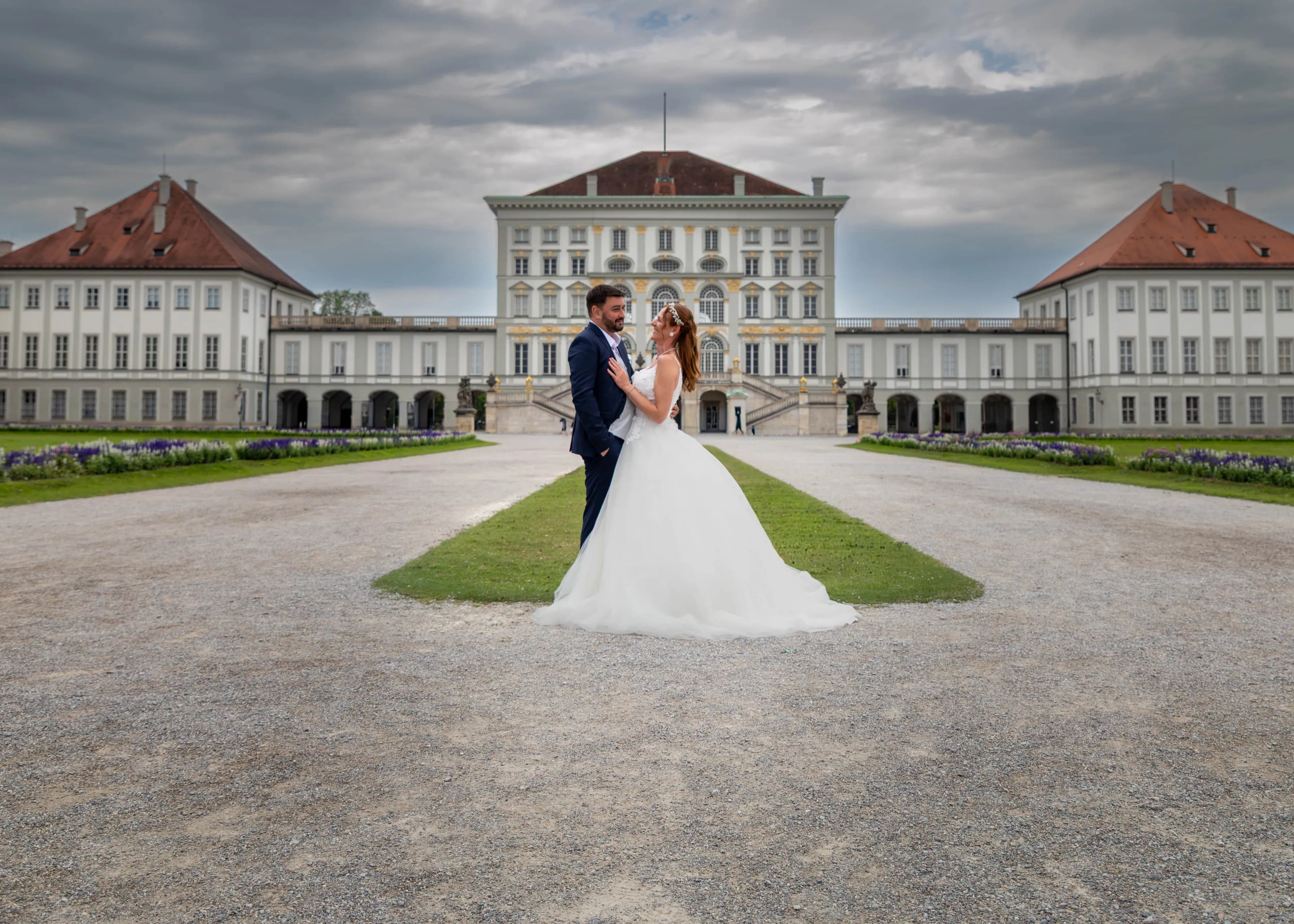 Brautpaar vor dem Schloss Nymphenburg in München, hinter ihnen die weiten Flügel der Schlossanlage unter Wolkenhimmel