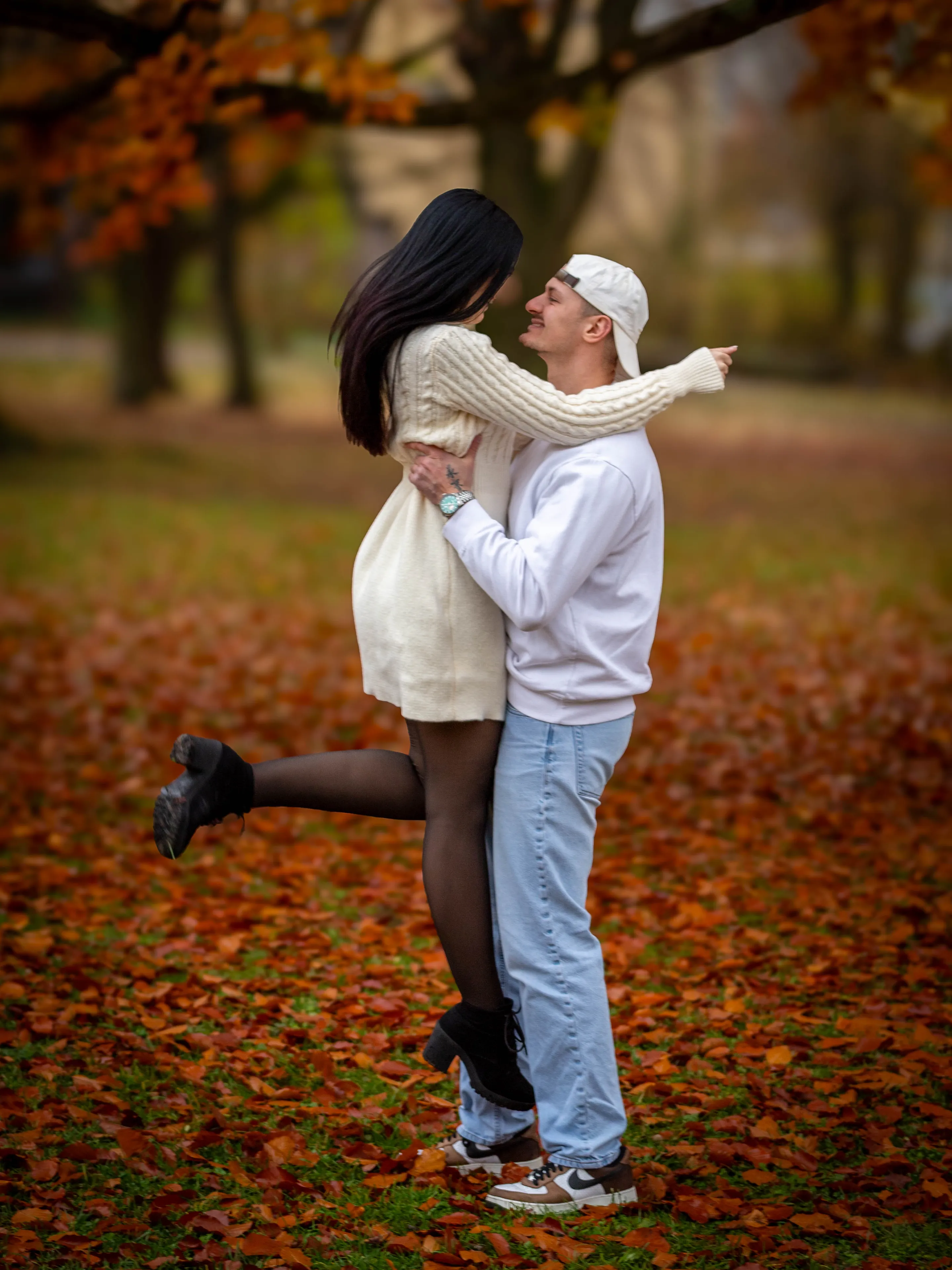 Ein junger Mann hebt seine Freundin lachend hoch, unter ihnen liegt rotes Herbstlaub